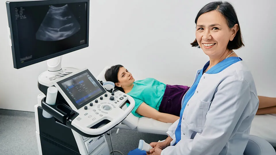 A sonographer smiles at the camera while her patient lies on a bed next to the scanning equipment A sonographer smiles at the camera while her patient lies on a bed next to the scanning equipment