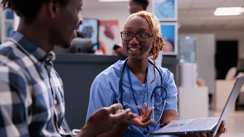 Medical assistant in scrubs discussing intake with patient, representing job duties and clinical work environment. Medical assistant in scrubs discussing intake with patient, representing job duties and clinical work environment.