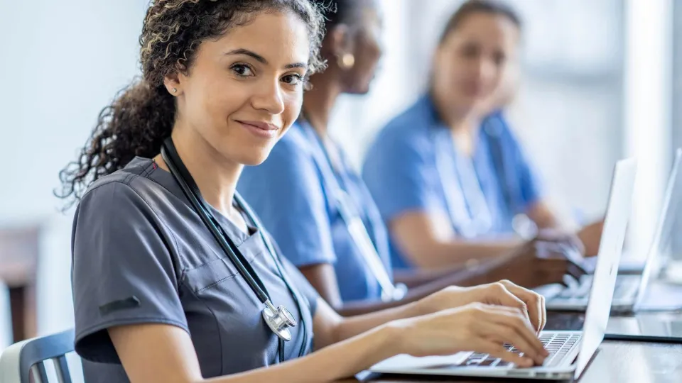 Smiling medical assistant student with stethoscope using laptop in class, representing medical assistant certification training. Smiling medical assistant student with stethoscope using laptop in class, representing medical assistant certification training.