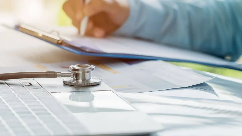 Person writing on a clipboard next to a laptop and stethoscope, representing health services administration responsibilities.