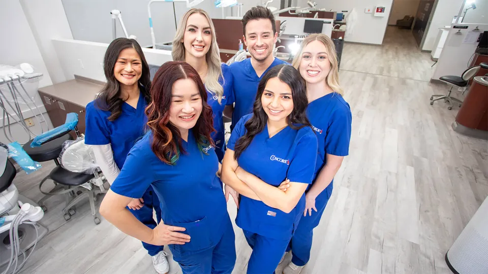 A group of smiling Concorde dental hygiene alumni stand together in blue scrubs within a Concorde dental training facility after completing their dental hygienist requirements.