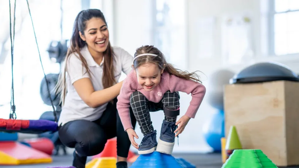 A young child balances on a large blue therapy ball while being guided through stability exercises by a pediatric physical therapist assistant.
