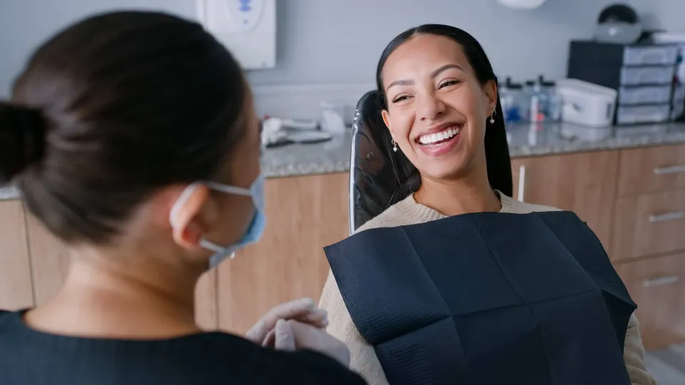 A smiling dental hygienist wearing a face mask and gloves demonstrates excellent communication skills while speaking with a happy patient in a dental chair.