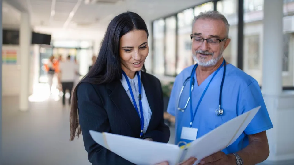 A female healthcare administrator in a black suit reviews medical documents in a binder with a senior male doctor in blue scrubs. A female healthcare administrator in a black suit reviews medical documents in a binder with a senior male doctor in blue scrubs.