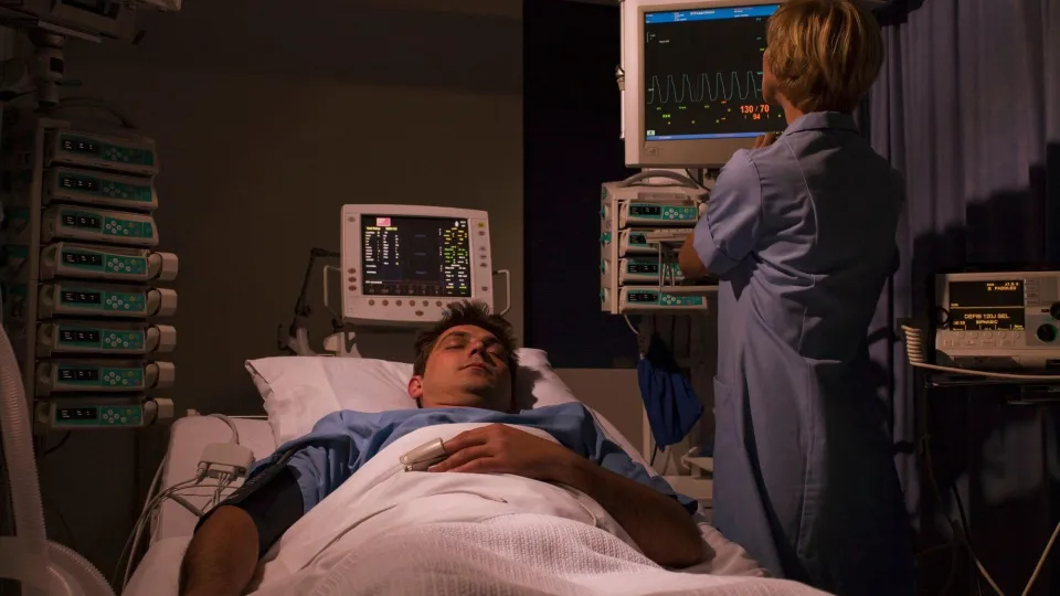A sleep technologist monitors real-time physiological data and vital signs on multiple screens while a patient sleeps in a darkened room.