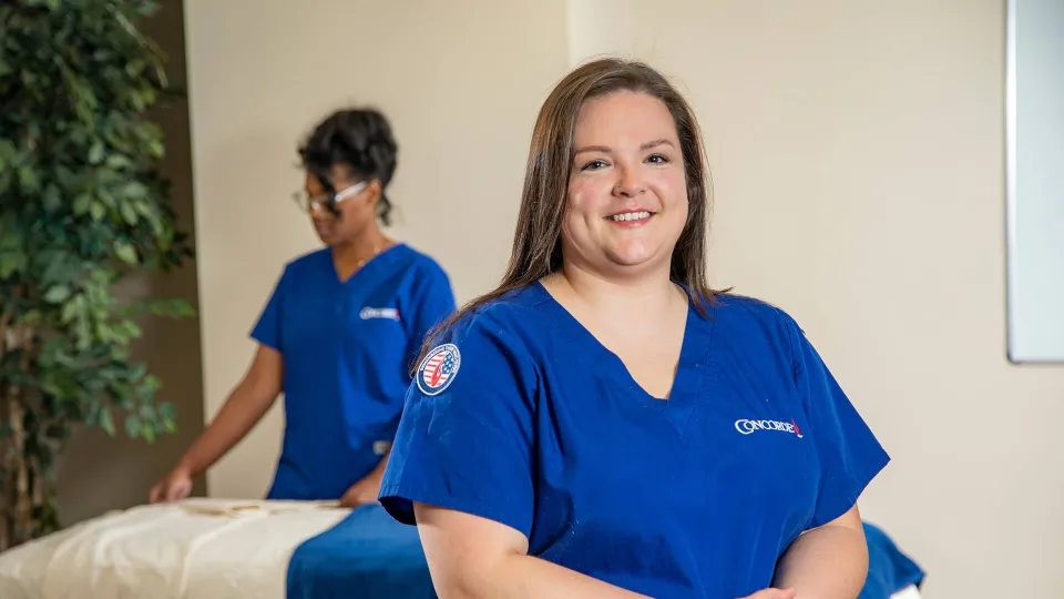 A smiling Concorde massage therapy student wearing blue scrubs with a military-themed patch stands in a healthcare training lab, illustrating how to use military tuition assistance for career training.