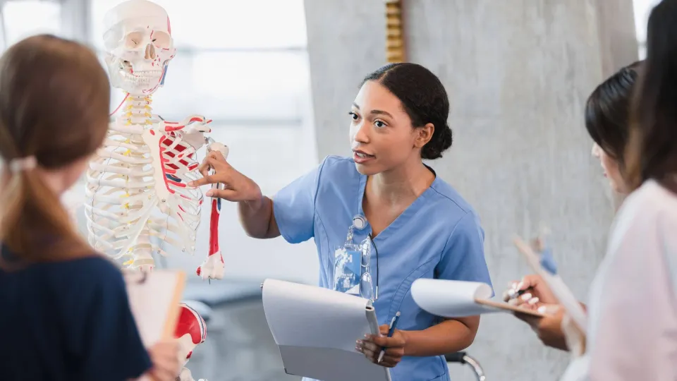 A female nurse educator uses a human skeleton model to teach clinical anatomy to a group of nursing students.
