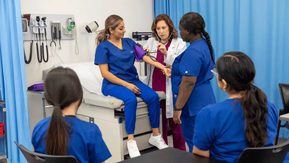 A healthcare professional and educator demonstrates how to take a patient's blood pressure using a student, while other students in the class watch