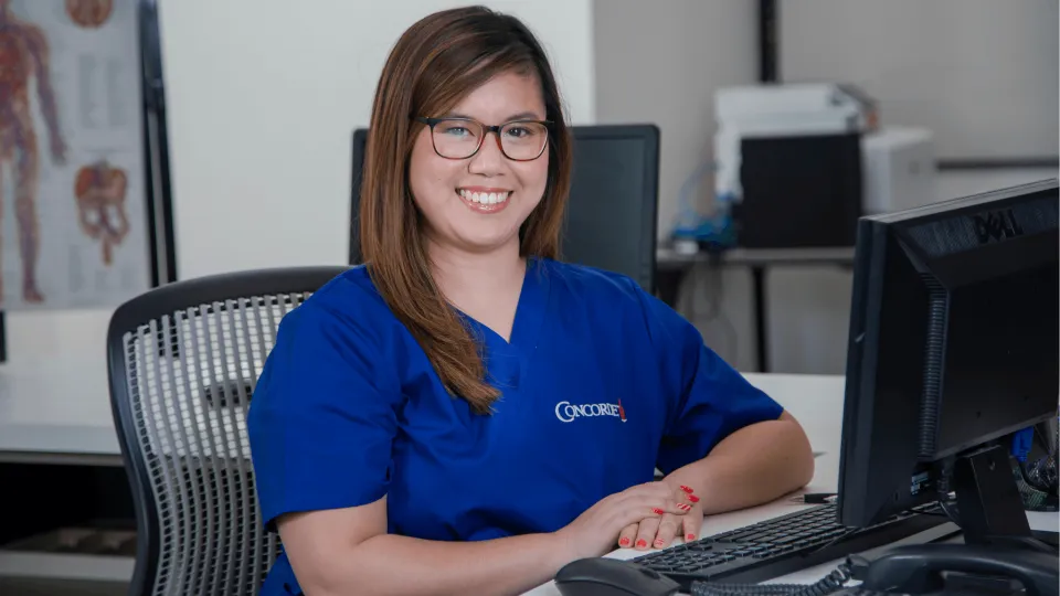 female medical office adminstrator in blue scrubs sits at computer and smiles for camera