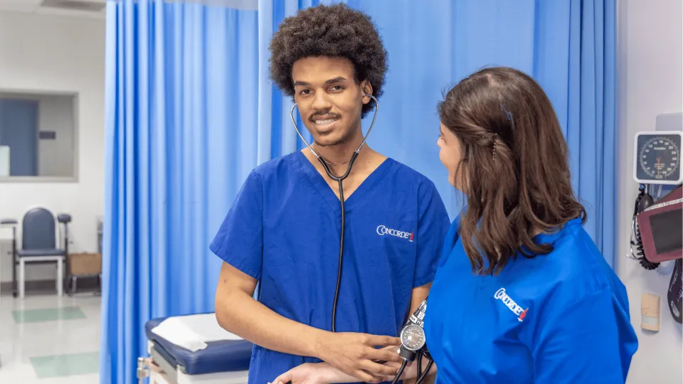 A male medical assistant wearing blue scrubs and stethescope works with patient and smiles for camera